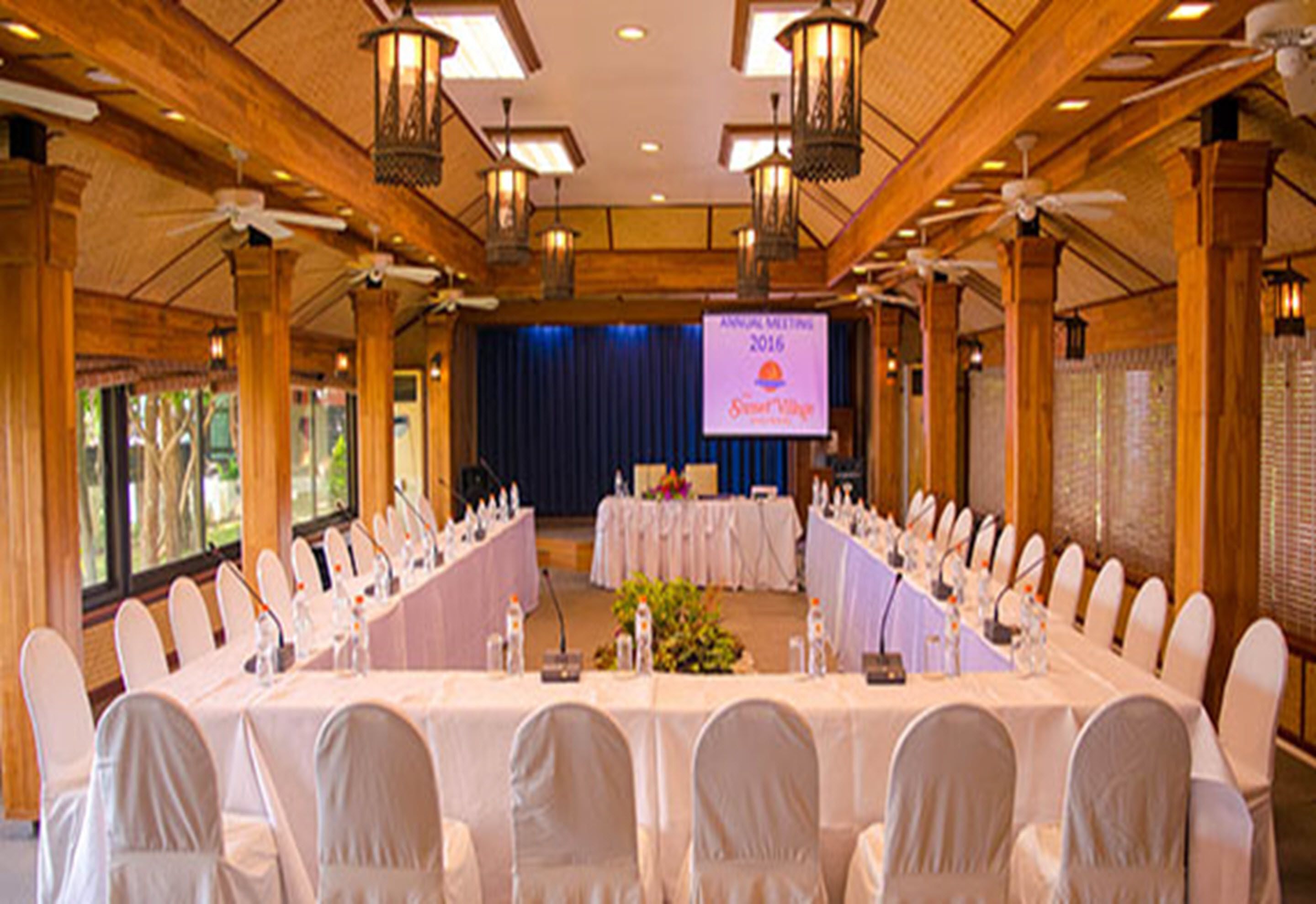 Un salón de conferencias con una larga mesa, sillas blancas y un escenario con una pantalla.