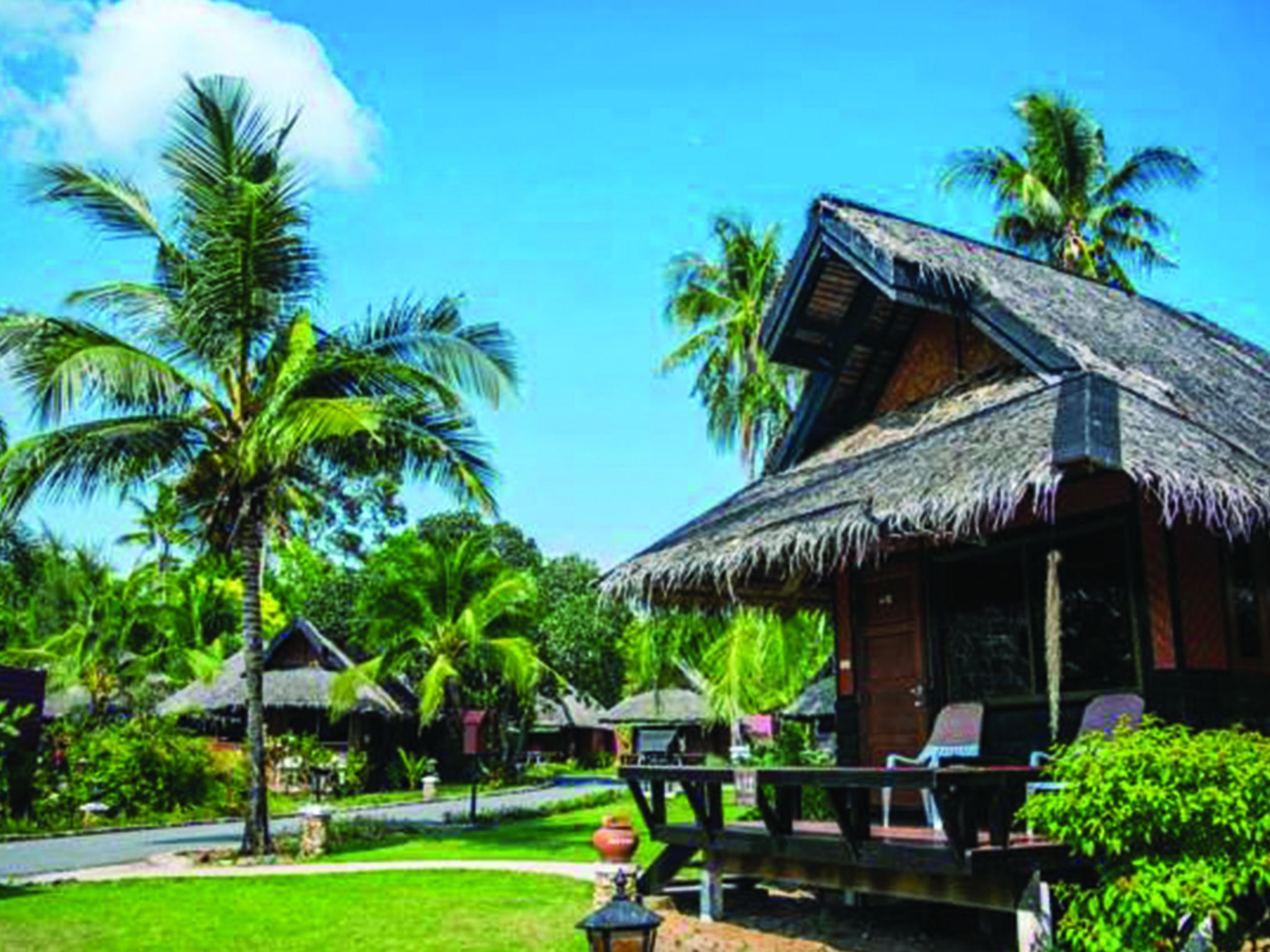 A thatched-roof bungalow with a porch, surrounded by lush greenery and palm trees.