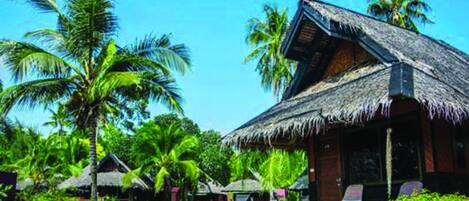 A thatched-roof bungalow with a porch, surrounded by lush greenery and palm trees.