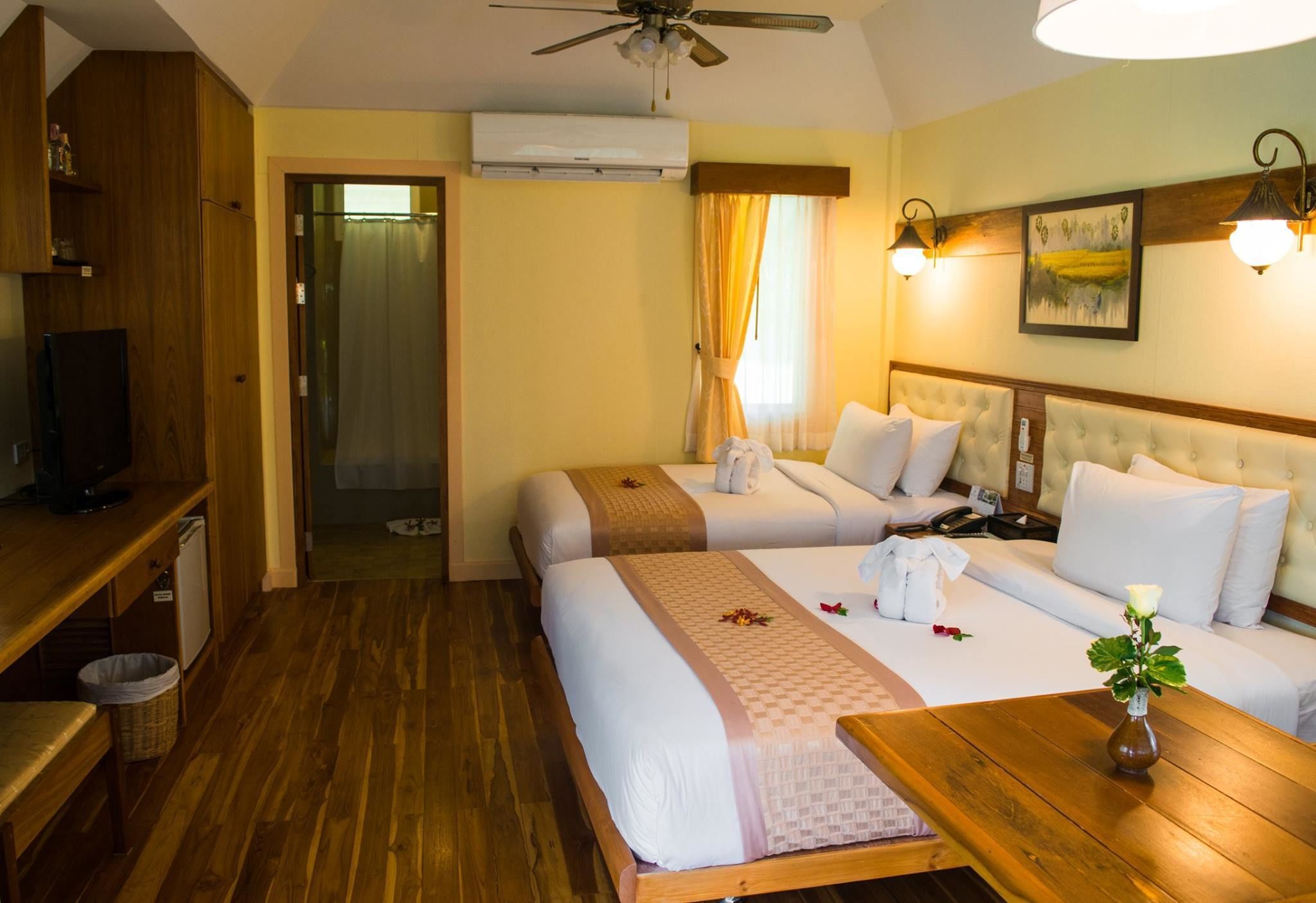 Habitación de hotel con dos camas, un escritorio de madera, un televisor y un ventilador de techo.