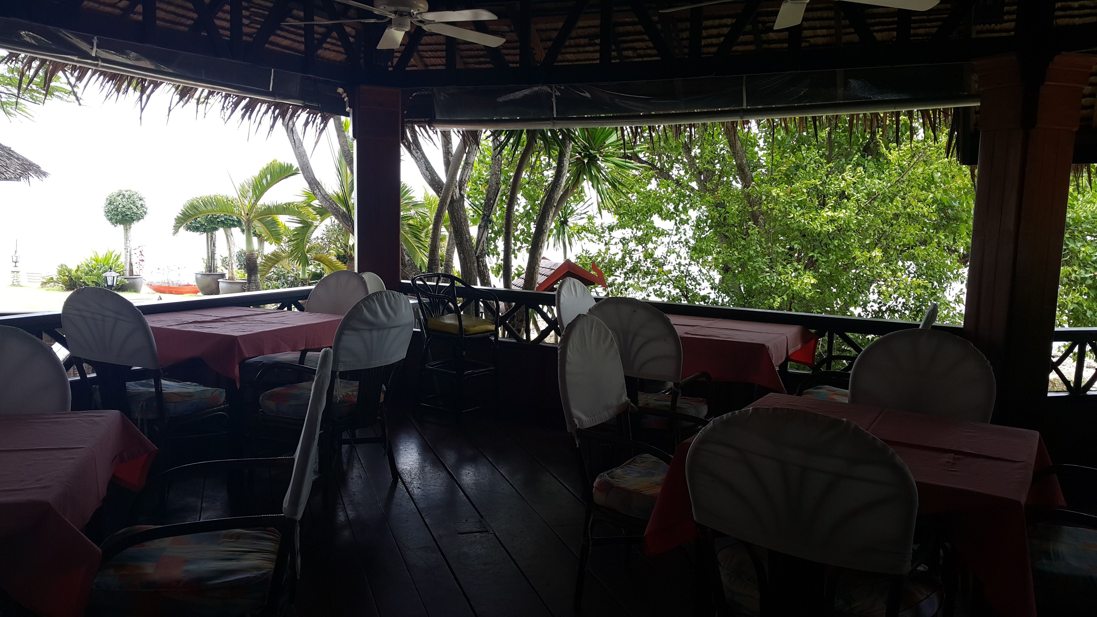 Un restaurante al aire libre con mesas y sillas, cubierto por un techo y rodeado de árboles.
