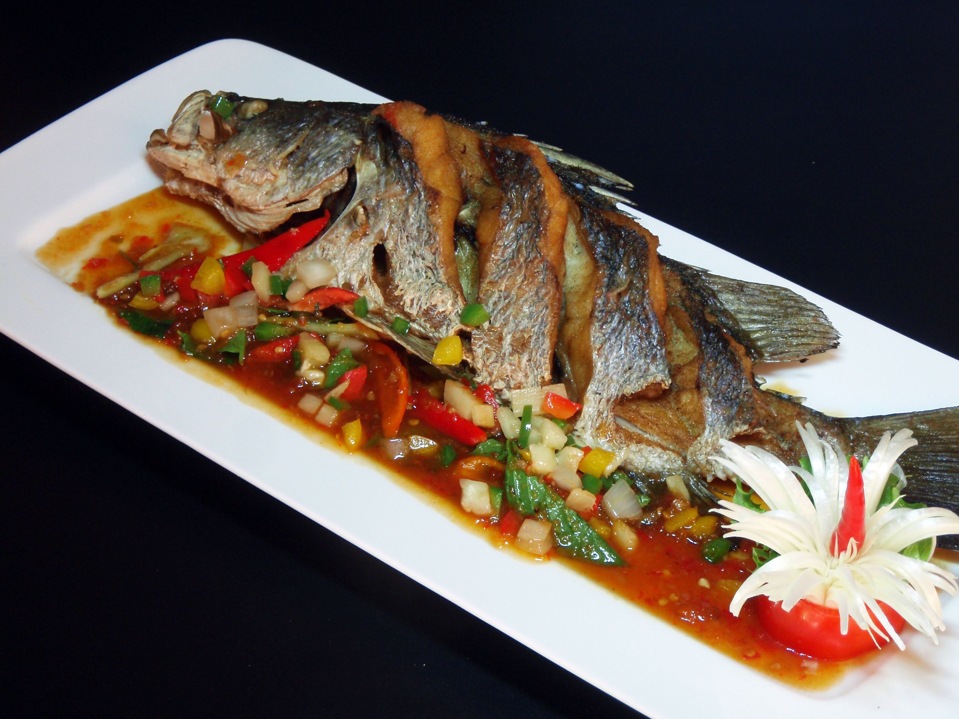 A white rectangular plate with a grilled fish, a red sauce, and a garnish of white and red flowers.