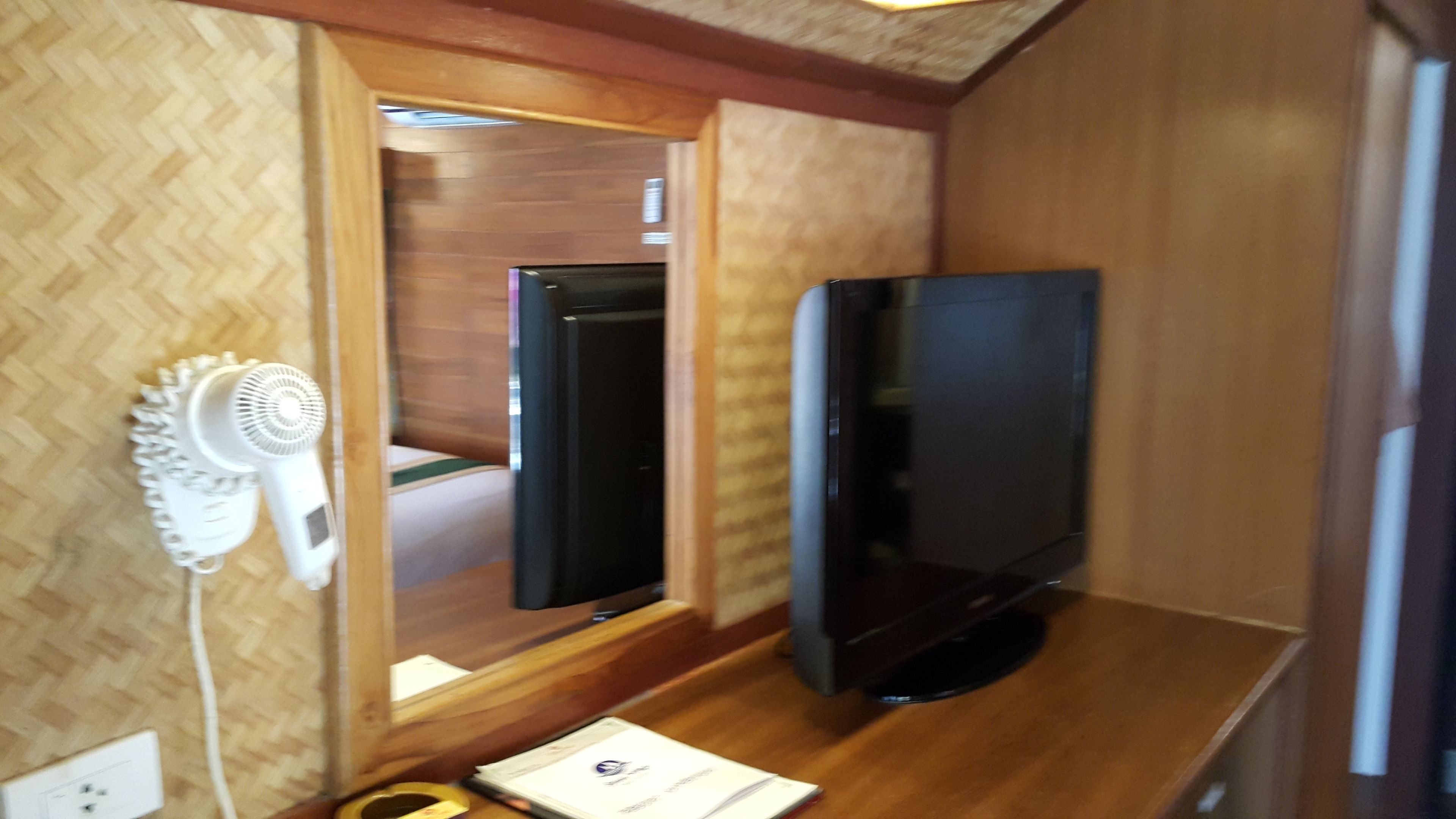 A compact room with a wooden floor, a flat-screen TV mounted on the wall, a white hairdryer, and a small table with a book.