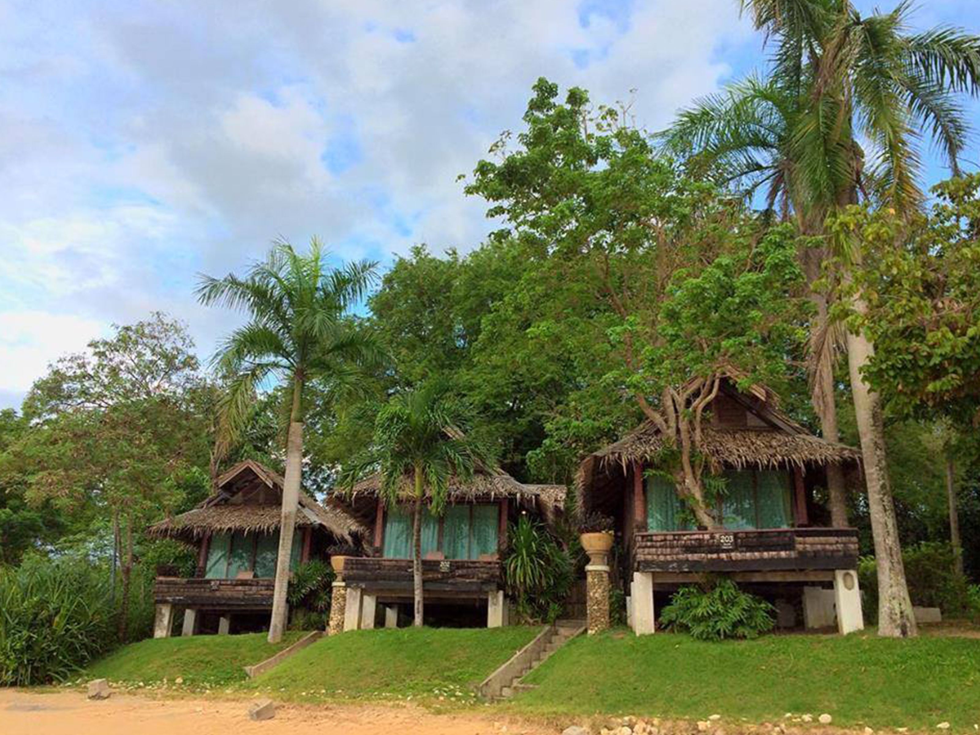 A row of thatched-roof cottages with wooden balconies, surrounded by lush greenery and palm trees.