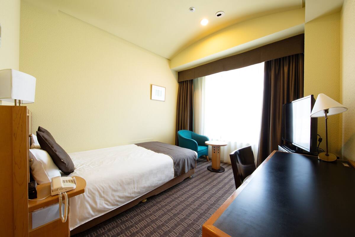 Ein Hotelzimmer mit Bett, Schreibtisch, Stuhl, Nachttisch, Lampe, Telefon und einem Fenster mit Vorhängen.