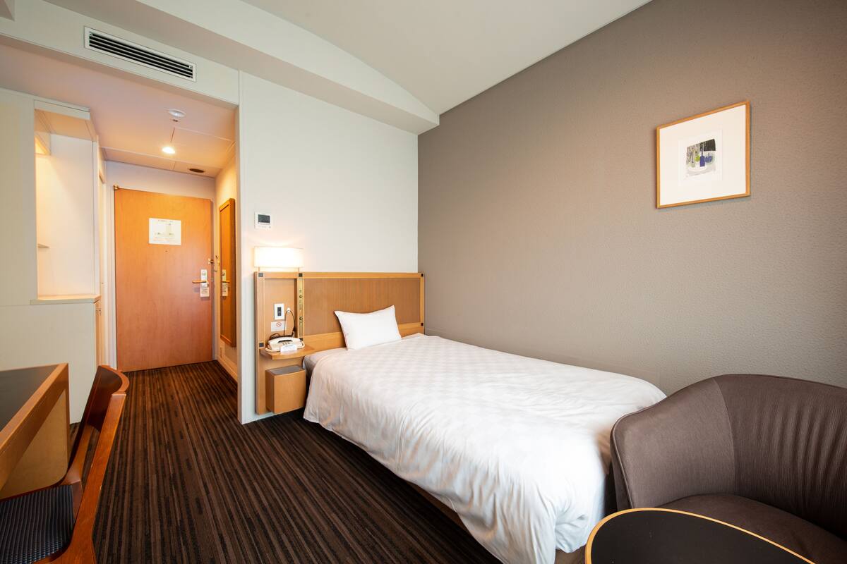 Ein Hotelzimmer mit einem Einzelbett, einem Schreibtisch, einem Stuhl, einem Nachttisch und einem gerahmten Bild an der Wand.
