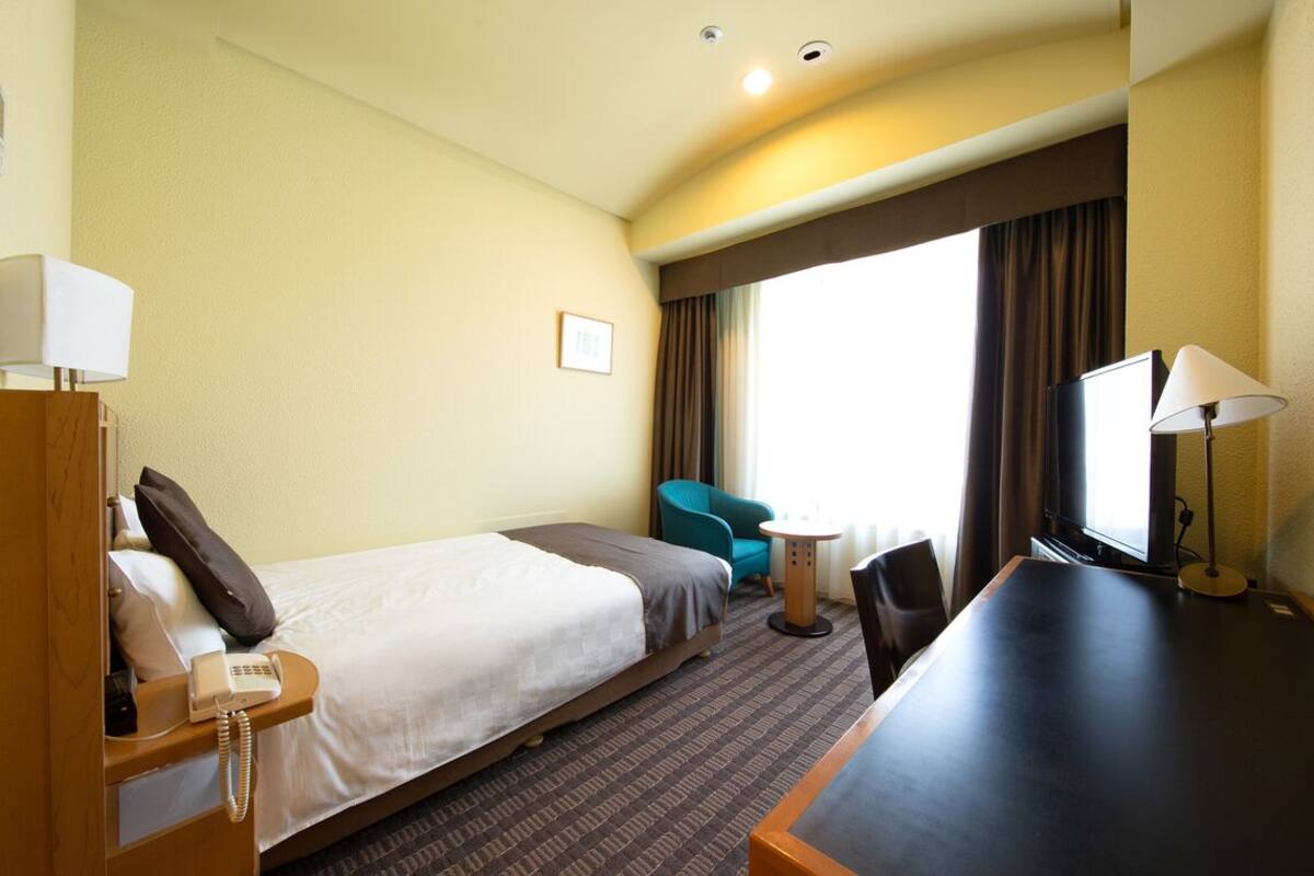 Ein Hotelzimmer mit Bett, Schreibtisch, Stuhl, Telefon, Lampe, Fernseher und einem Fenster mit Vorhängen.