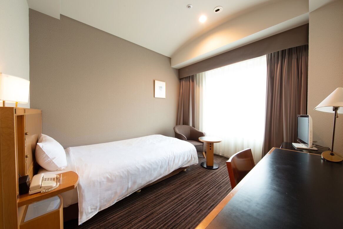 Habitación de hotel con cama, escritorio, silla, mesita, televisor y ventana con cortinas.