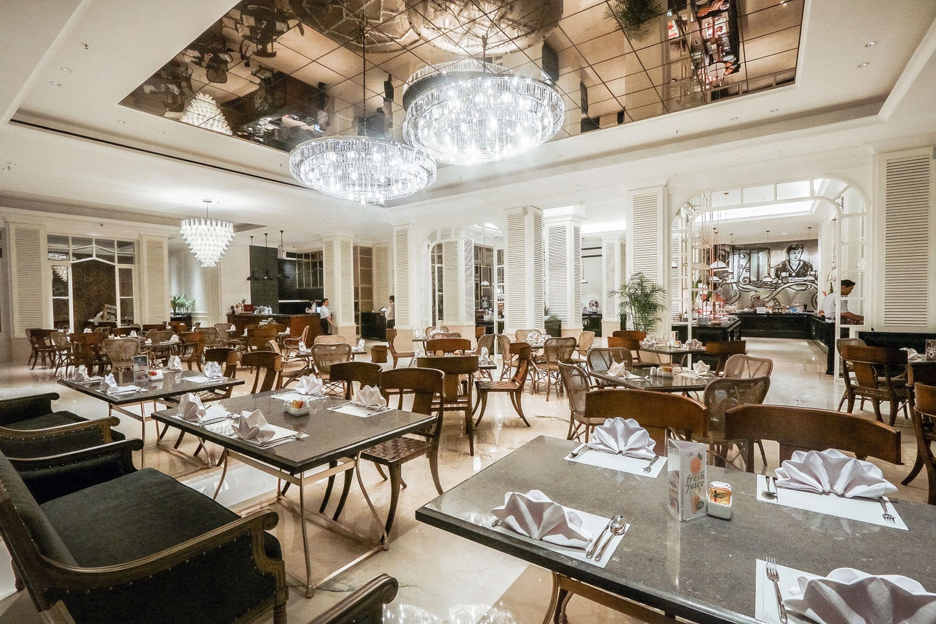 A spacious dining area with multiple tables set for service, elegant chandeliers, and a high ceiling with a skylight.