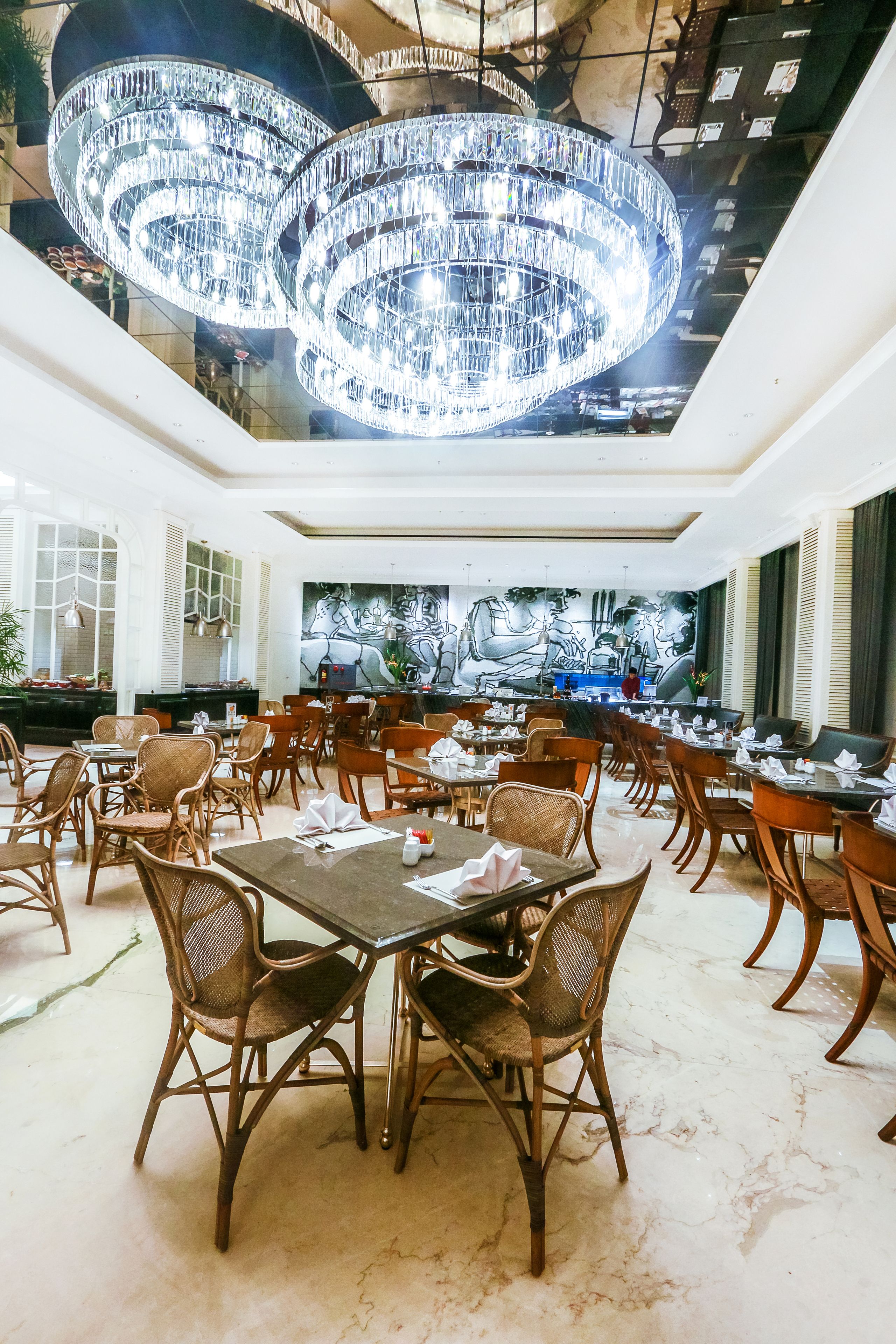 A spacious dining area with high ceilings, ornate chandeliers, and a large mural on the back wall.