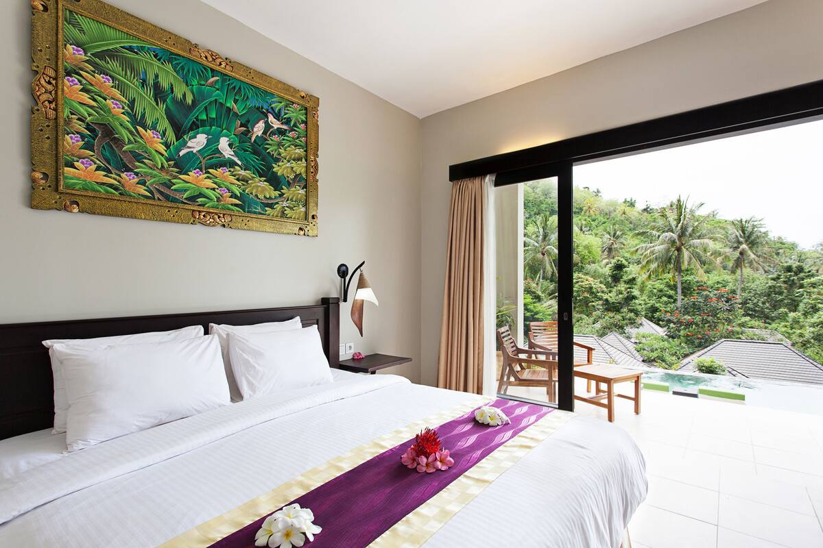 Ein Hotelzimmer mit einem großen Bett, einem Gemälde, einem Balkon mit Blick auf Grünflächen und einem Tisch mit Stühlen im Freien.
