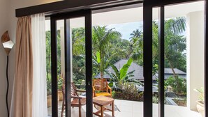 Una stanza con una porta scorrevole in vetro che dà su un patio con mobili in legno e vista su un giardino tropicale.