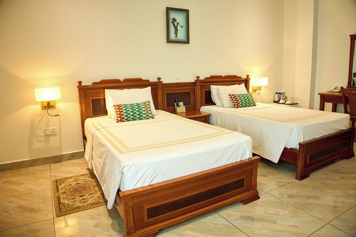 A hotel room with two wooden beds, each with a white bedspread and pillows, a bedside table with a lamp, and a framed picture on the wall.