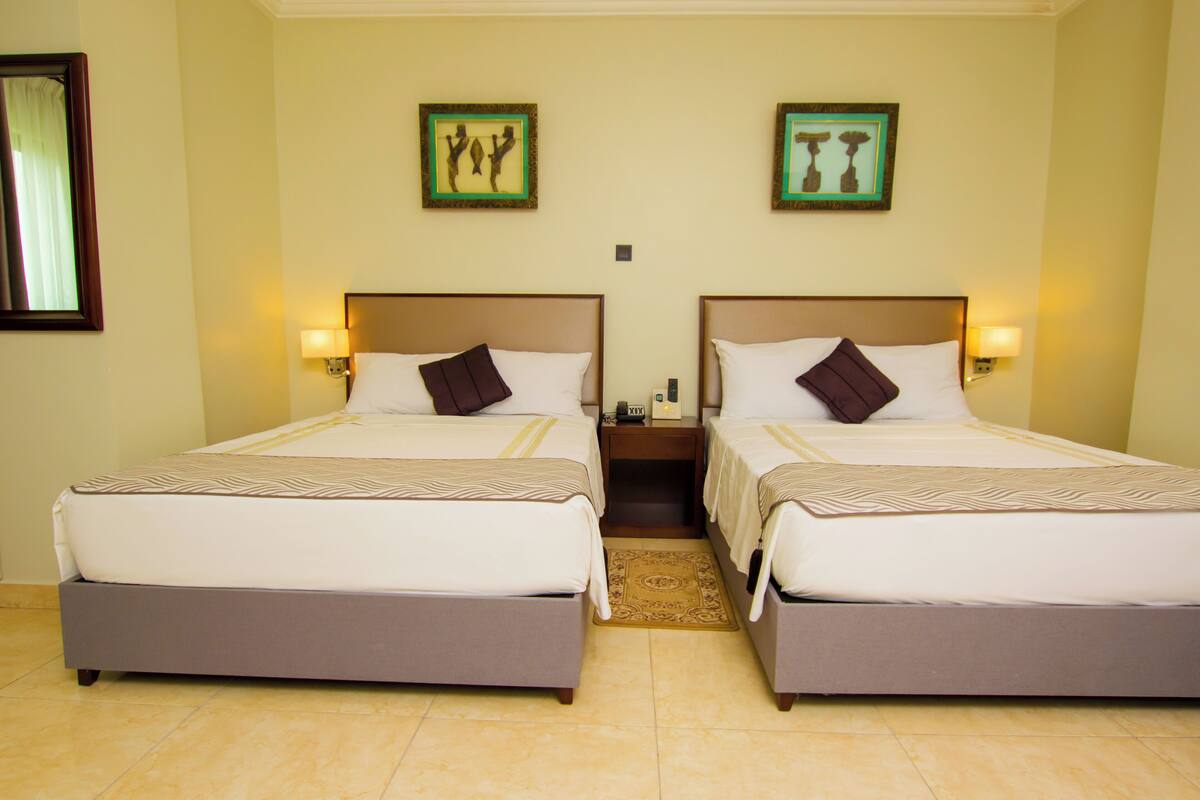 A hotel room with two beds, each with a headboard, bedside lamps, and a small table between the beds.