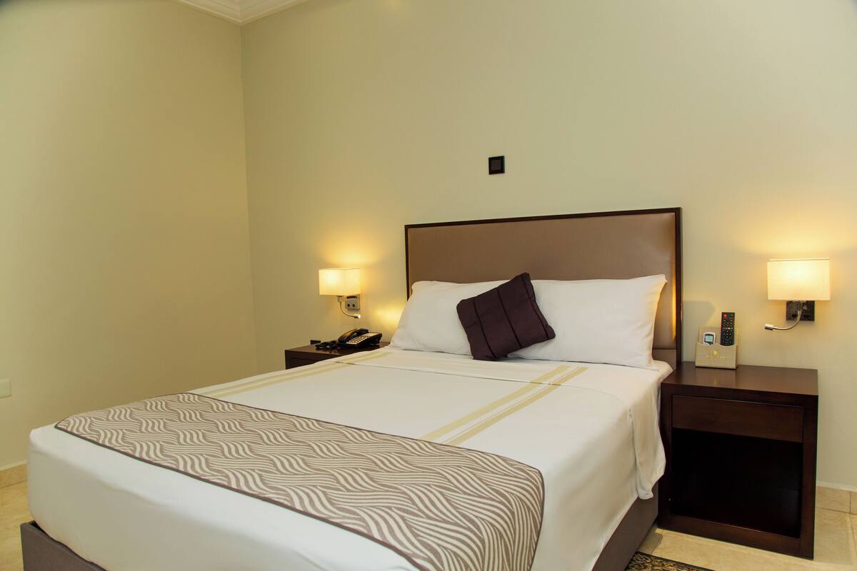 A hotel room with a large bed, two bedside tables with lamps, a telephone, and a wall-mounted control panel.