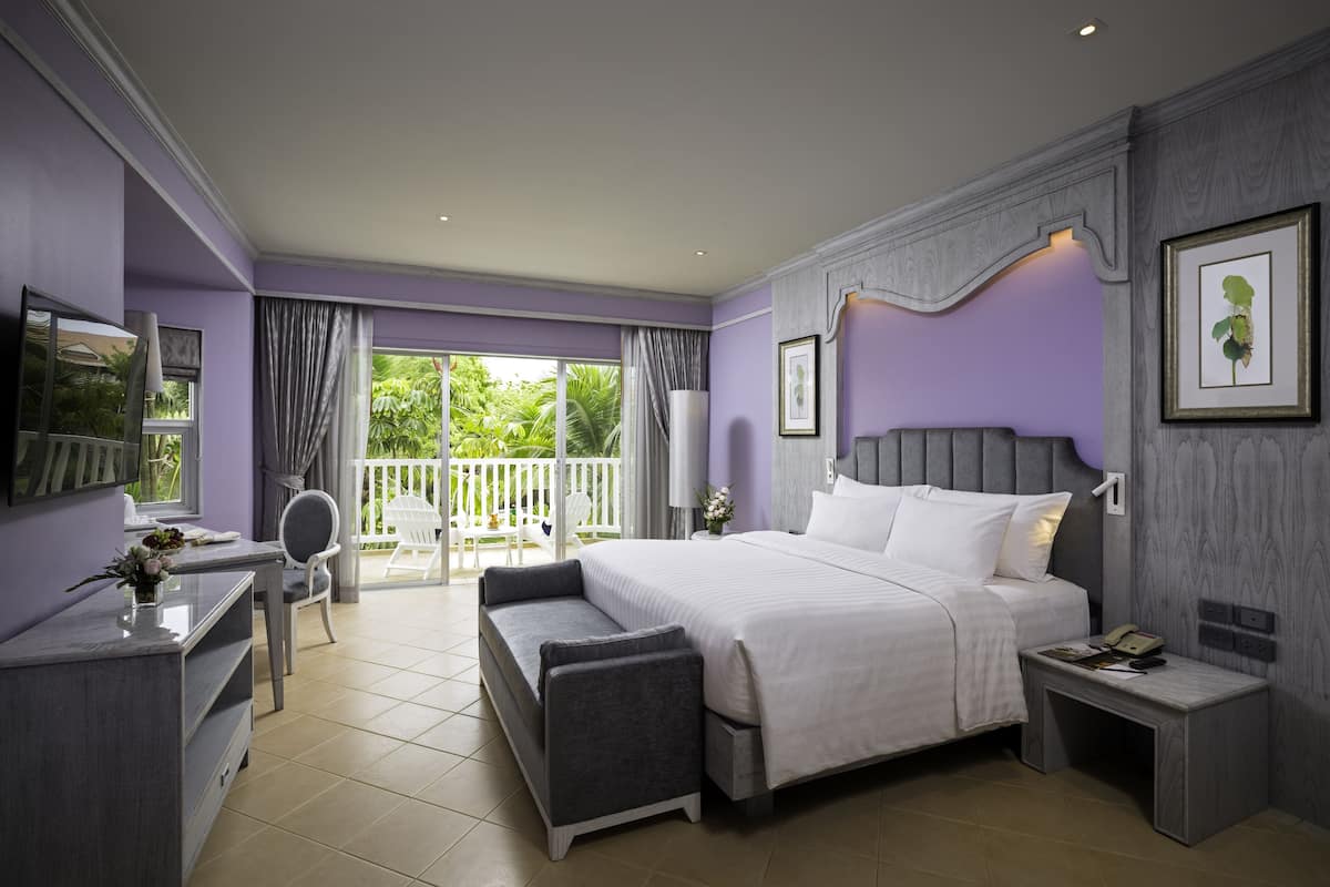 A modern bedroom with a large bed, a desk, and a chair. There is a balcony with a view of greenery.