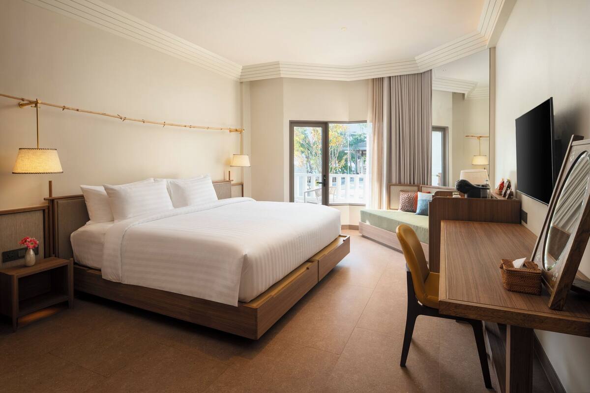 A hotel room with a large bed, bedside tables with lamps, a wooden desk, a chair, a flat-screen TV, and a balcony with a view.