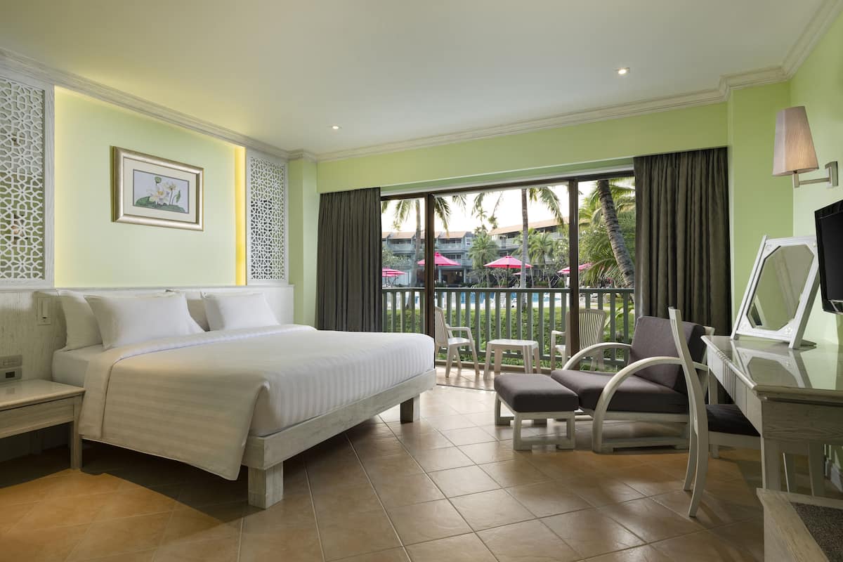 A hotel room with a bed, a desk with a chair, a TV, and a balcony with a view of palm trees and umbrellas.