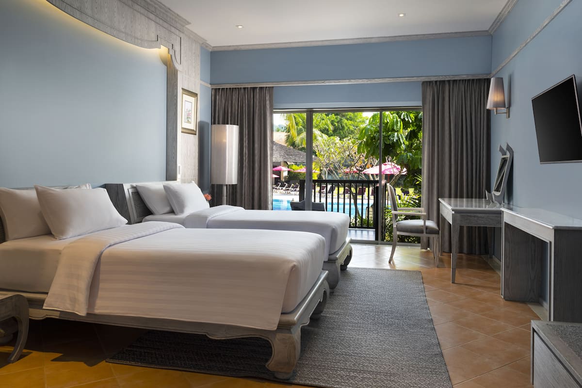 A hotel room with two beds, a desk, a TV, and a view of a pool and outdoor seating.