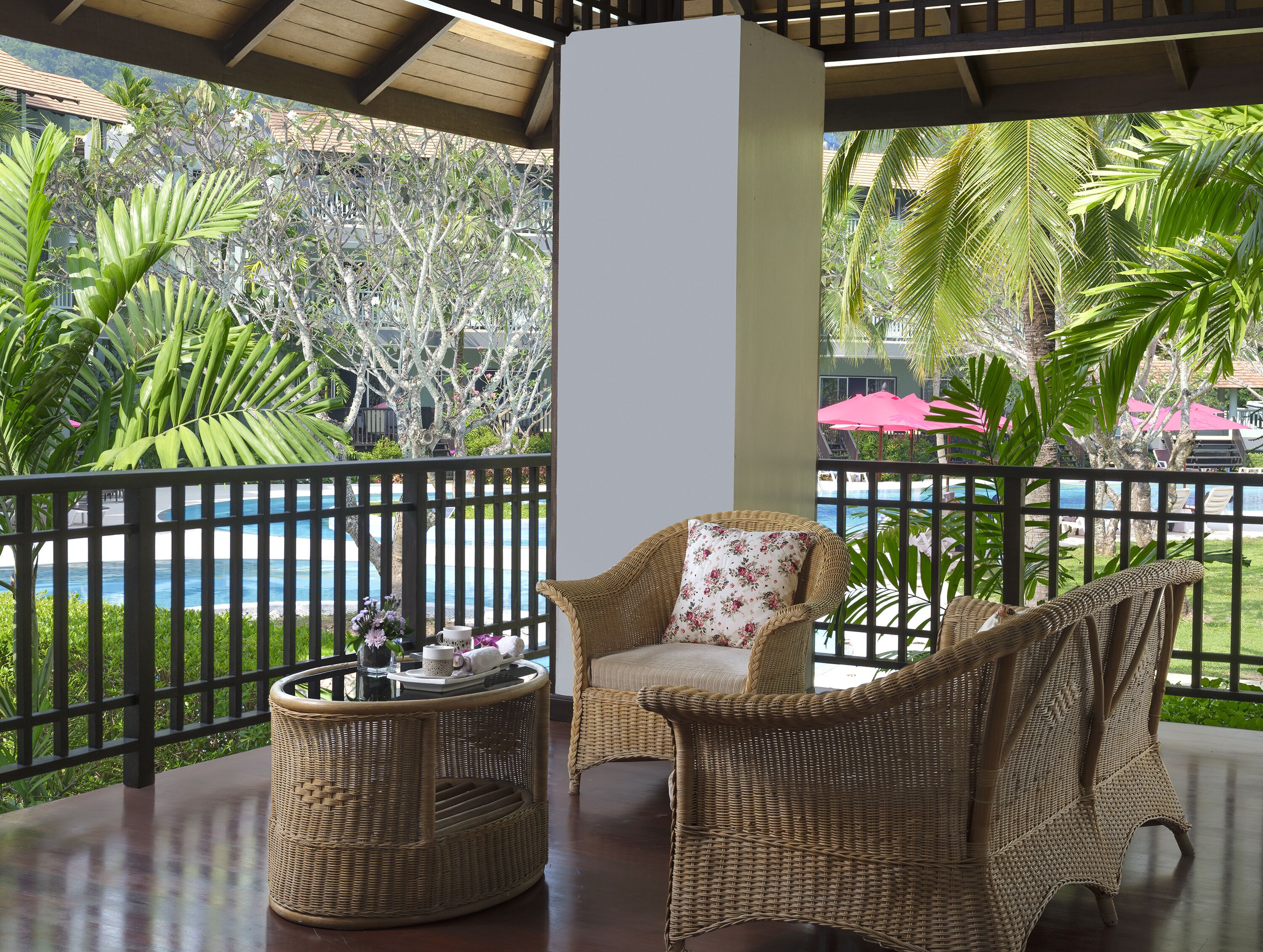Une chaise en osier avec un coussin à motifs floraux, un panier en osier et une vue sur une piscine et des plantes tropicales.