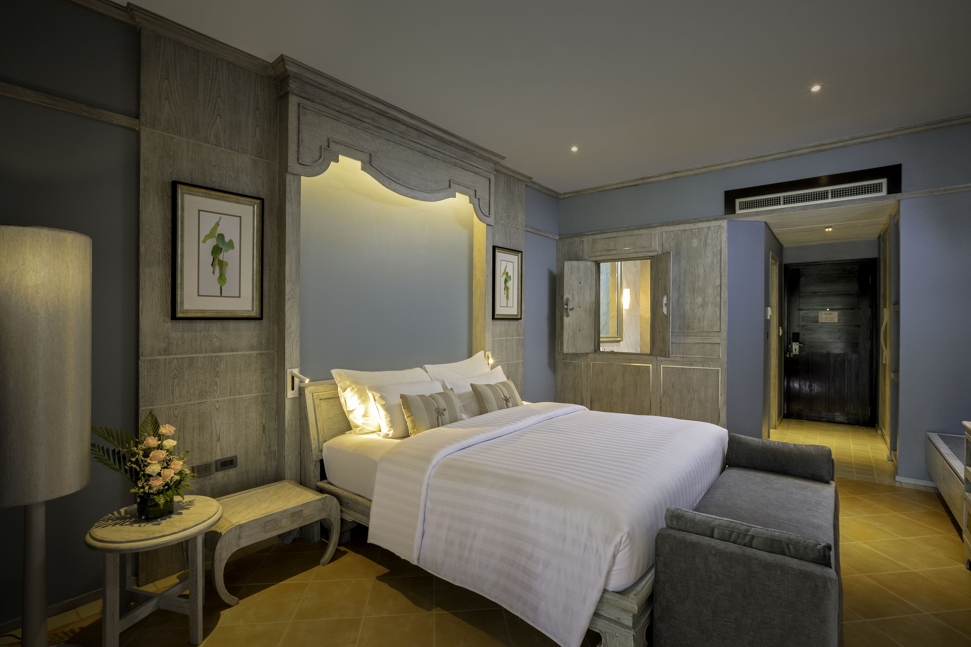 A bedroom with a large bed, a bedside table with a lamp, a bench, and a small table with flowers.