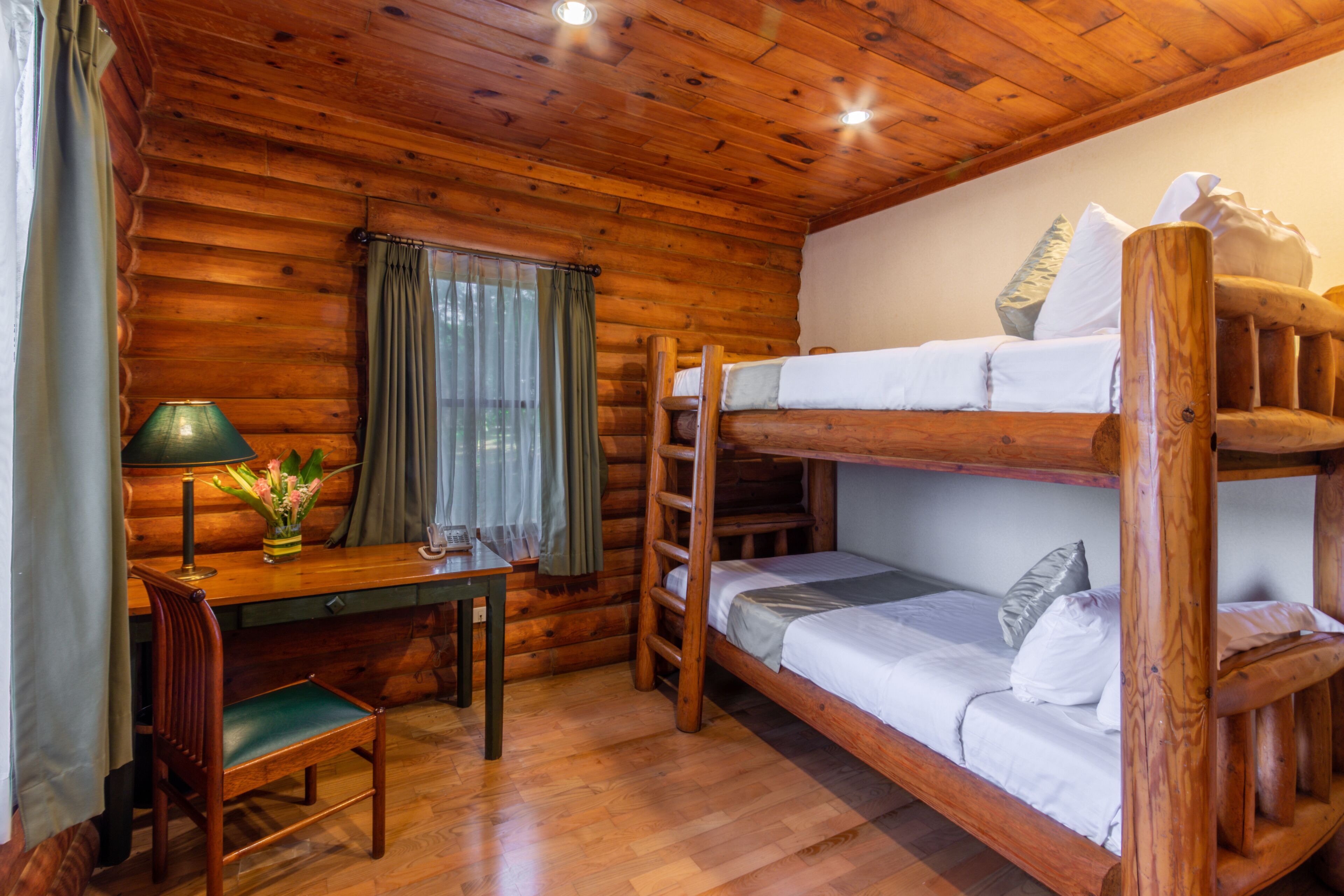 A cabin-style room with a bunk bed, a desk with a chair, a lamp, and a window with curtains.