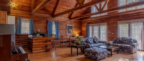 A log cabin interior with a piano, sofa set, dining table, and large windows.