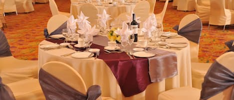 Un salón de banquetes con mesas redondas dispuestas para un evento formal, cada una con mantel blanco, cubertería y cristalería.