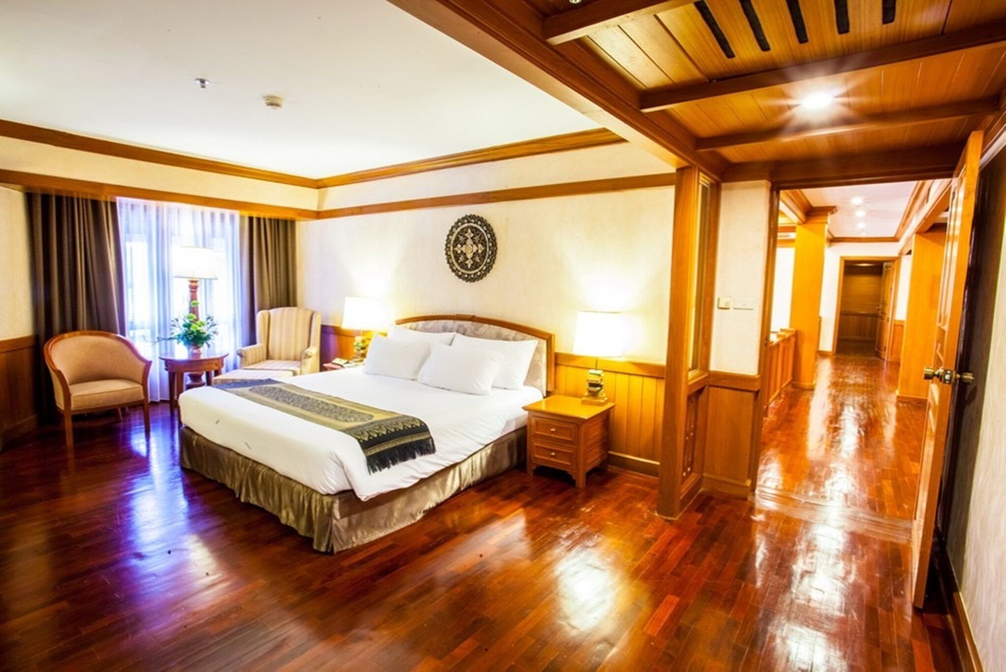 A hotel room with a large bed, wooden flooring, a desk, a chair, and a nightstand with a lamp.