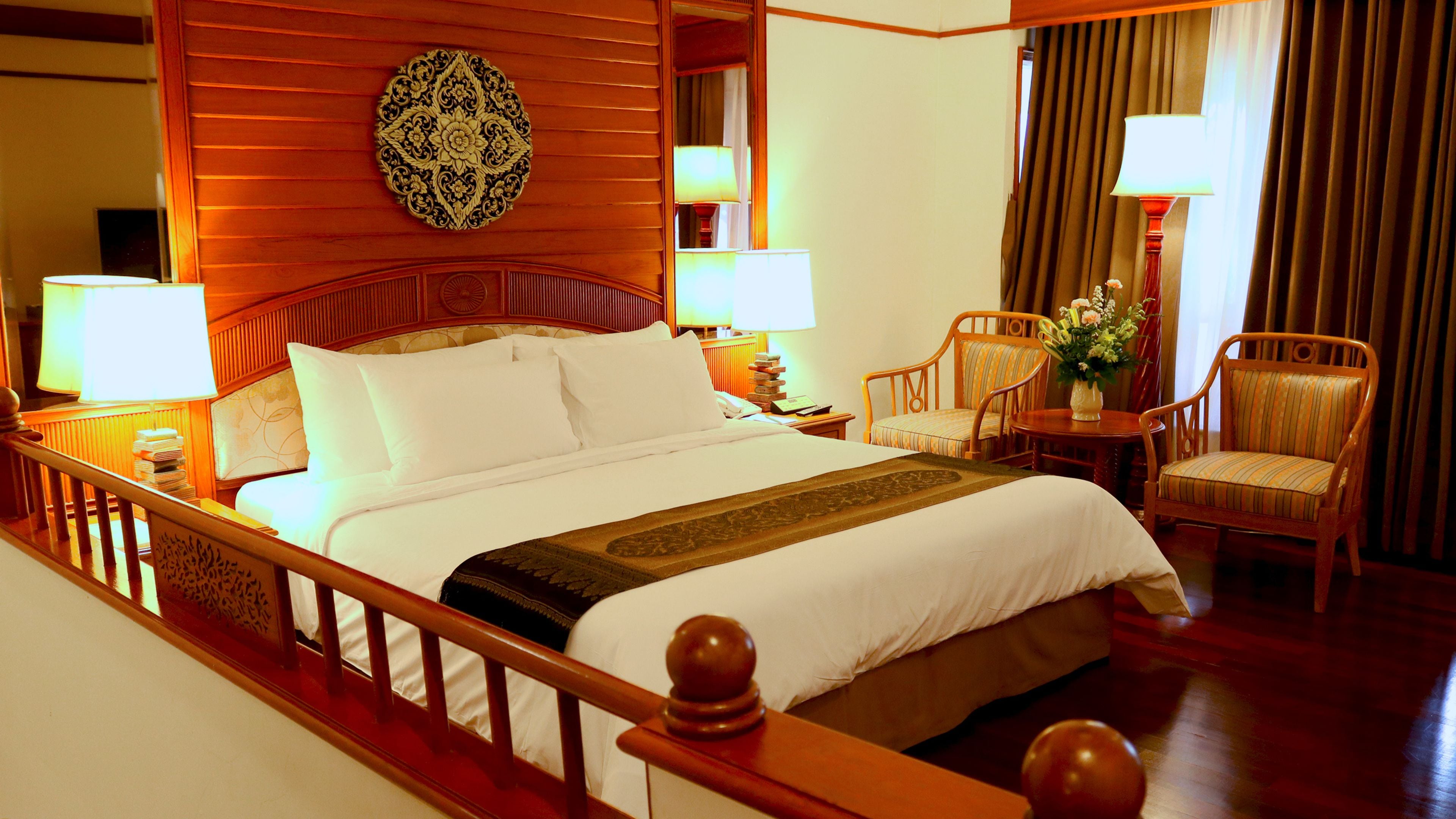 Habitación de hotel con una cama grande, dos mesitas de noche con lámparas, una silla de madera, una pequeña mesa con un florero y un adorno en la pared.
