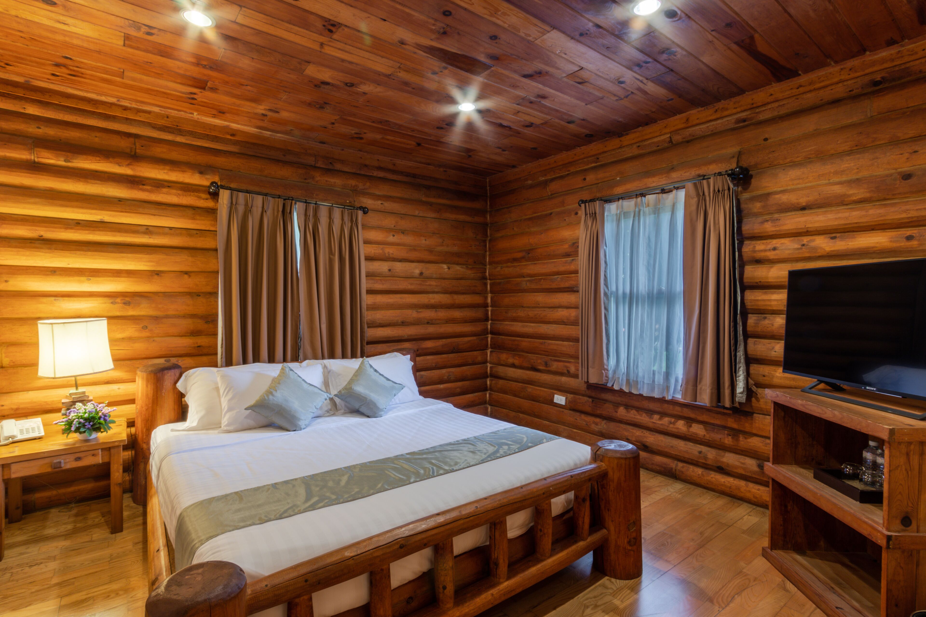 Een blokhutkamer met een houten bed, een televisie op een houten standaard, en een nachtkastje met een lamp en telefoon.