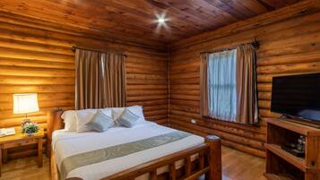 Een blokhutkamer met een houten bed, een televisie op een houten standaard, en een nachtkastje met een lamp en telefoon.