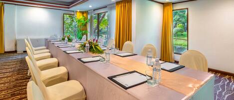 A conference room with a long table, chairs, and a view of greenery through large windows.