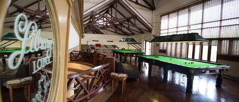 A spacious indoor pool area with multiple pool tables, wooden railings, and large windows allowing natural light.