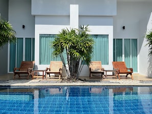 Zona de piscina con tumbonas de madera y una palmera, rodeada por un edificio blanco con ventanas grandes y cortinas verdes.