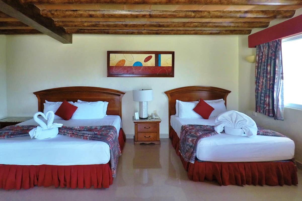 Habitación de hotel con dos camas, cada una con falda roja, ropa de cama blanca y una toalla con forma de cisne. Se observa un cabecero de madera, una mesita de noche con lámpara y un cuadro en la pared.