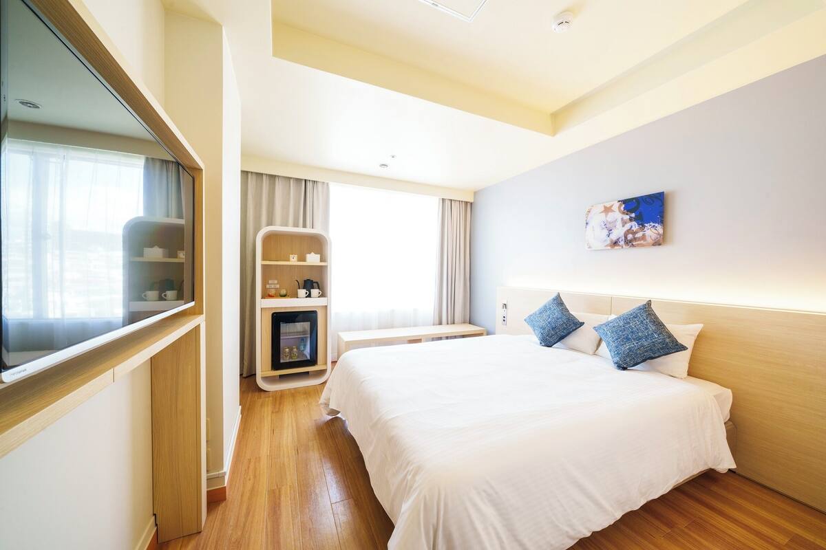 A hotel room with a large bed, a bedside table, a TV, a small seating area with a table and chairs, and a window with curtains.