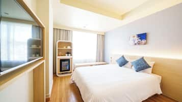 Habitación de hotel con una cama grande, una mesita de noche, televisor, un pequeño rincón de descanso con una mesa y sillas, y una ventana con cortinas.