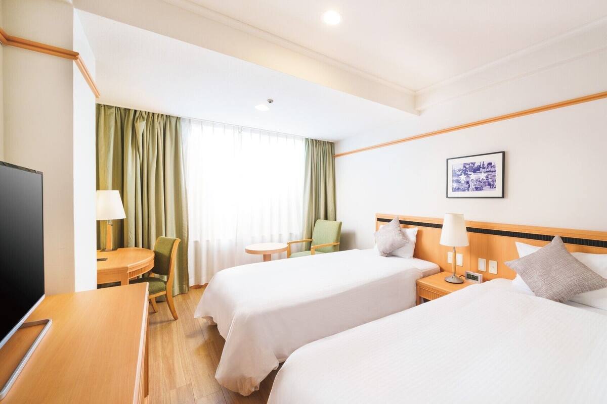 A hotel room with two beds, a desk, a chair, a TV, and a window with curtains.