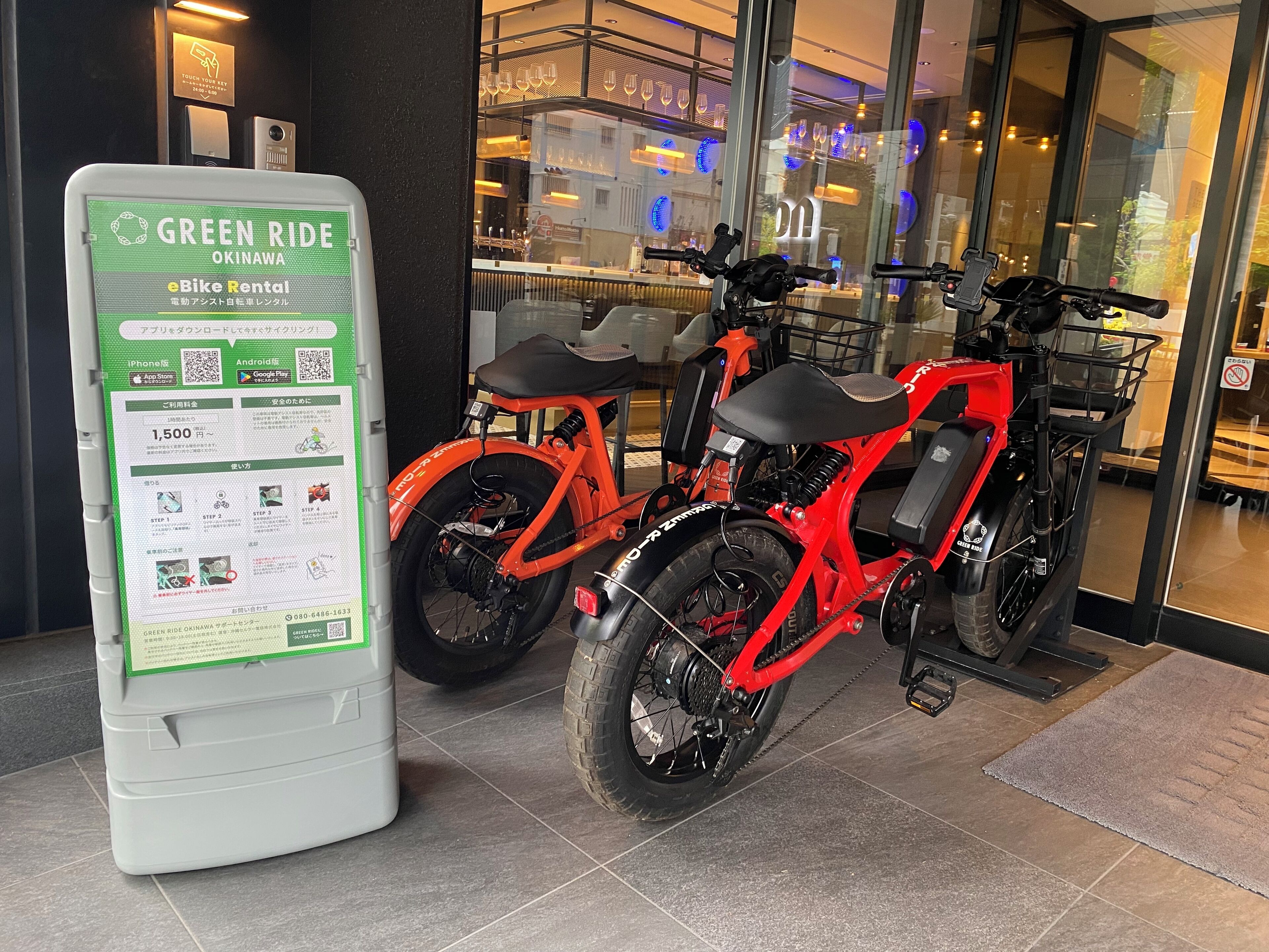 A green bike rental station with instructions and a QR code, two red eBikes, and a glass storefront with indoor seating.