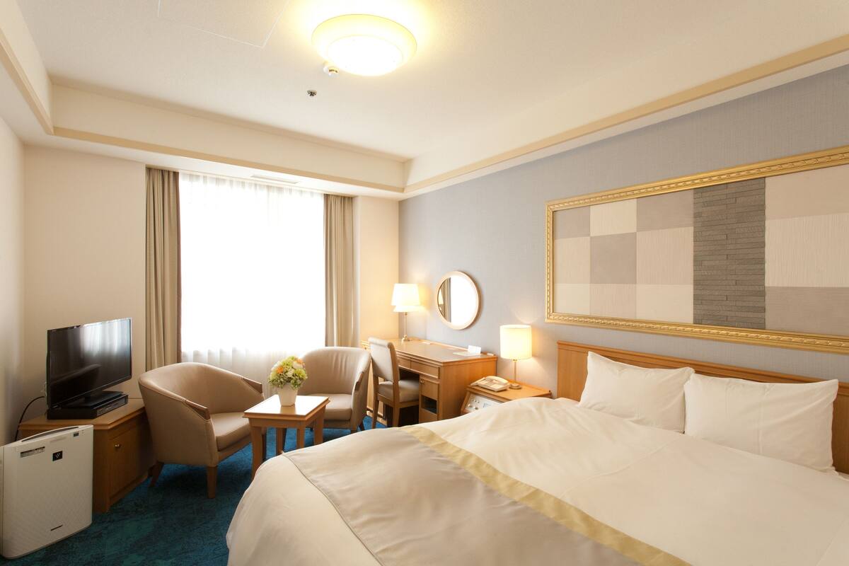 A hotel room with a large bed, a desk with a chair, a television, a desk lamp, and a large mirror on the wall.