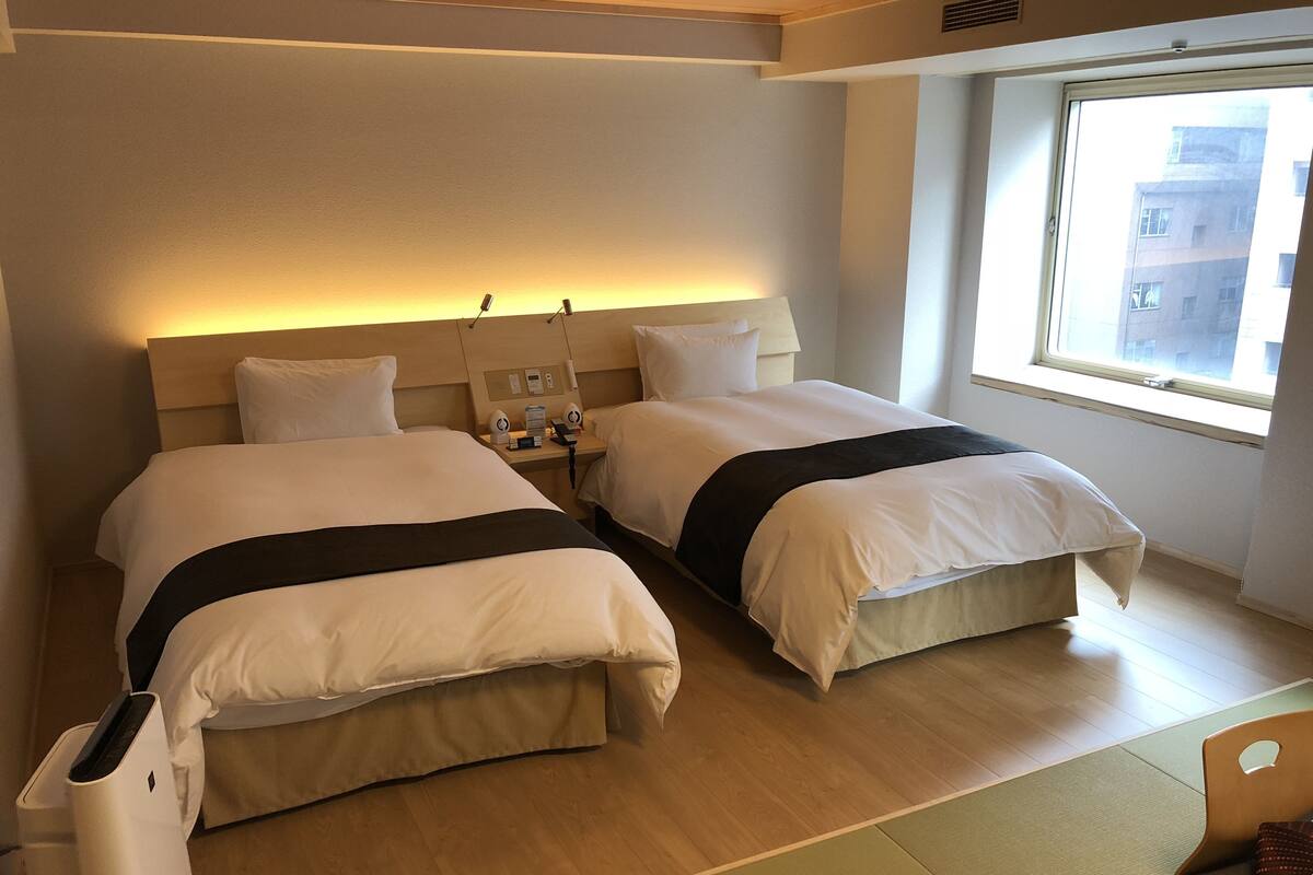A hotel room with two beds, a bedside table with a phone and a lamp, a window with a view of buildings, and a wooden floor.