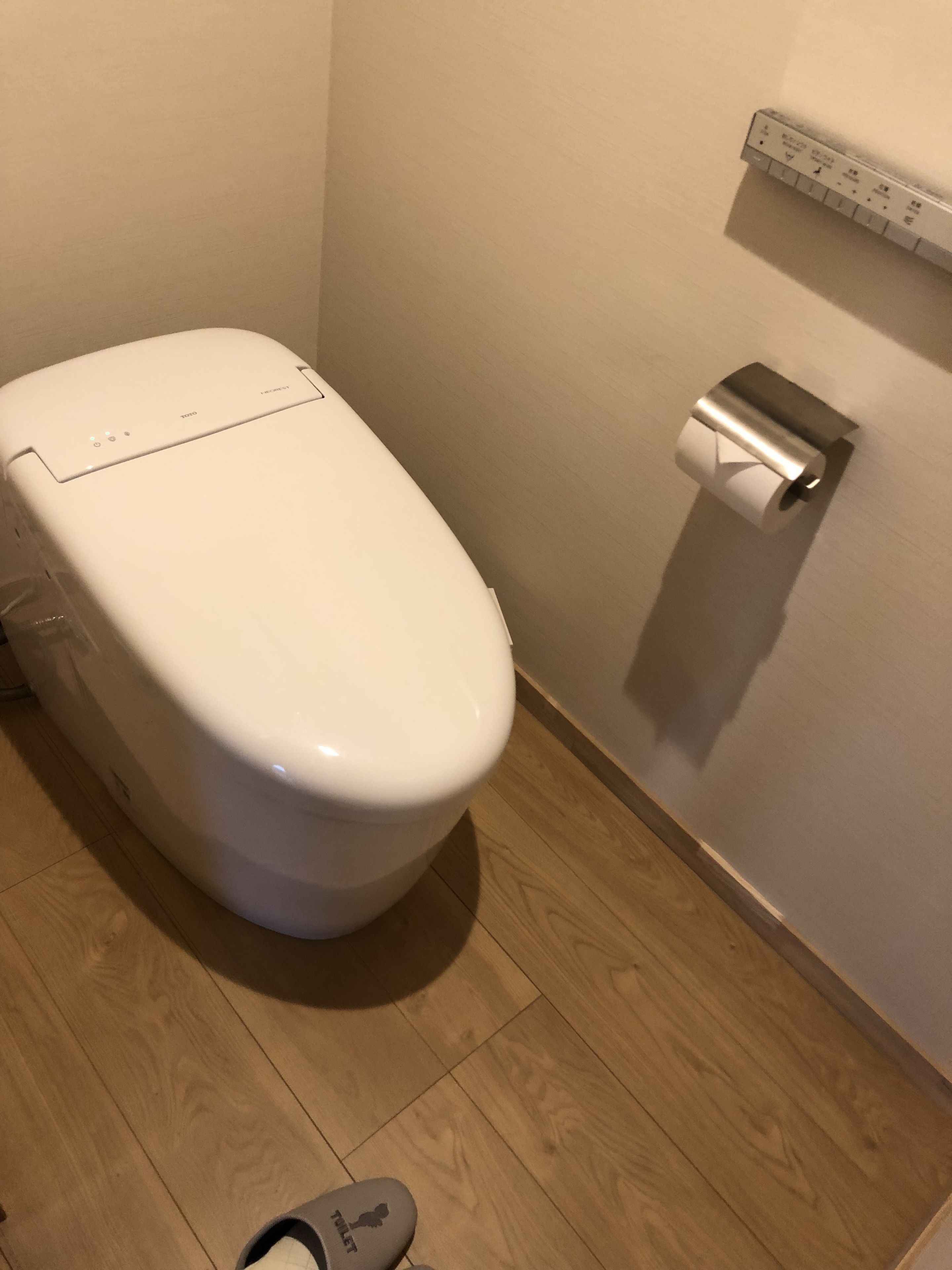 Des toilettes blanches avec une pédale au sol, un porte-rouleau de papier toilette et un plancher en bois.