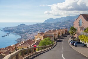 A coastal street with terracotta-roofed buildings, parked cars, and a view of the sea and distant hills. - Palheiro Village (Funchal)
