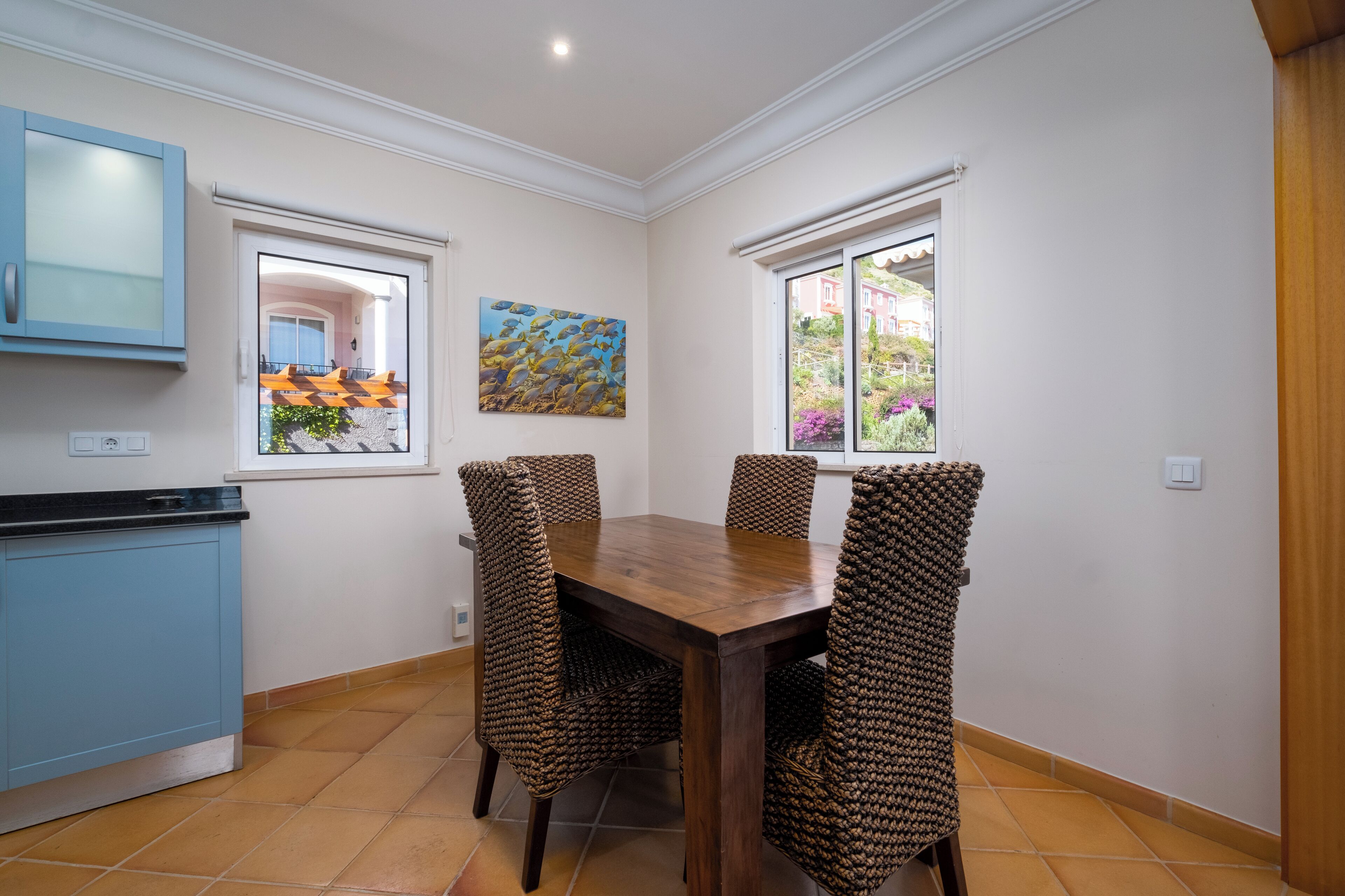 A dining area with a wooden table and four woven chairs, a blue cabinet, and a window with a view of greenery.