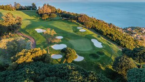 An aerial view of a golf course with sand bunkers, trees, and a coastline in the background.