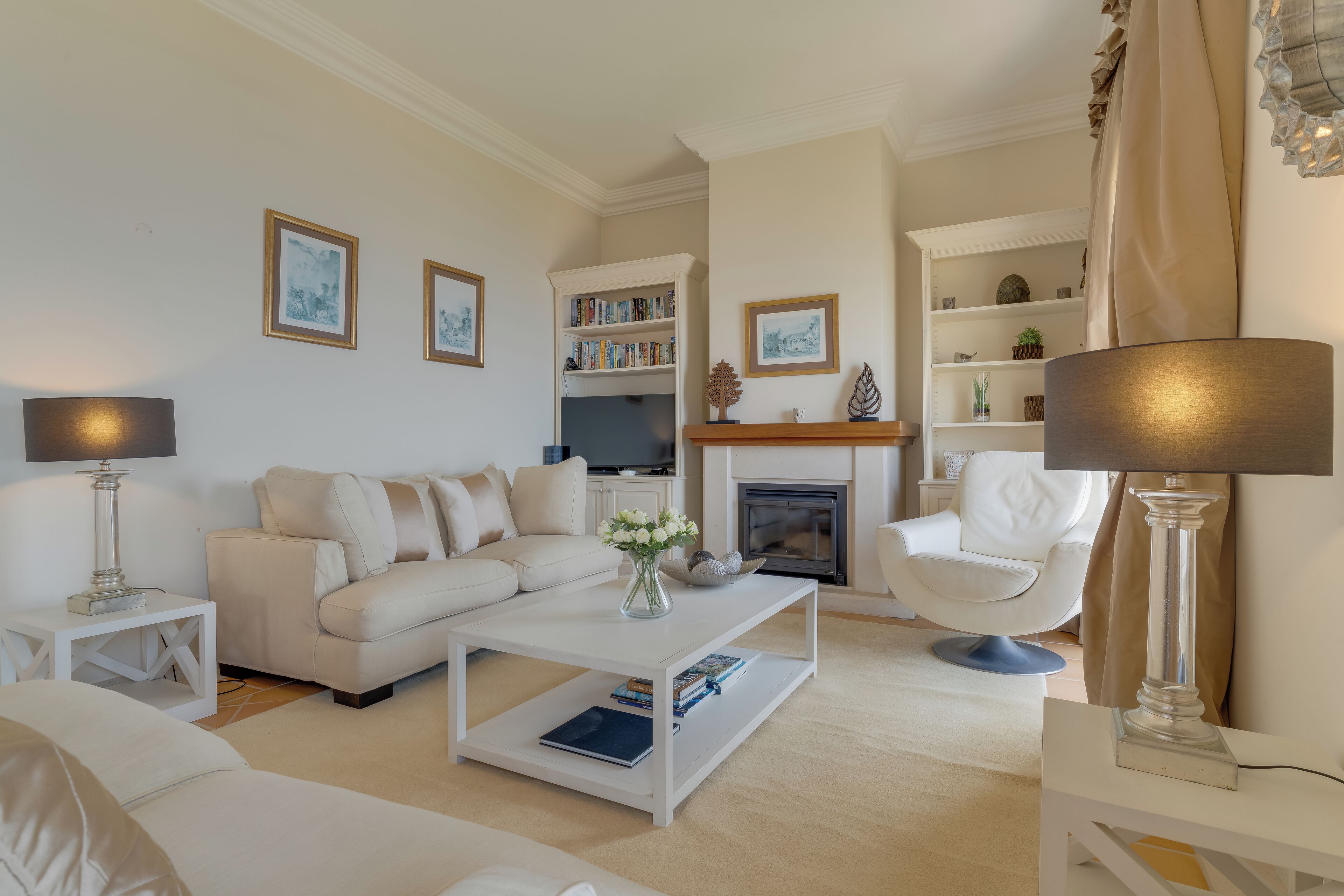 A modern living room with a sofa, coffee table, and armchairs. There is a fireplace, bookshelves, and a television.