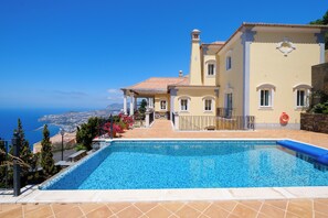 A poolside view of a Mediterranean-style villa with a clear view of the sea, a tiled patio, and a small outdoor seating area. - Palheiro Village (Funchal)