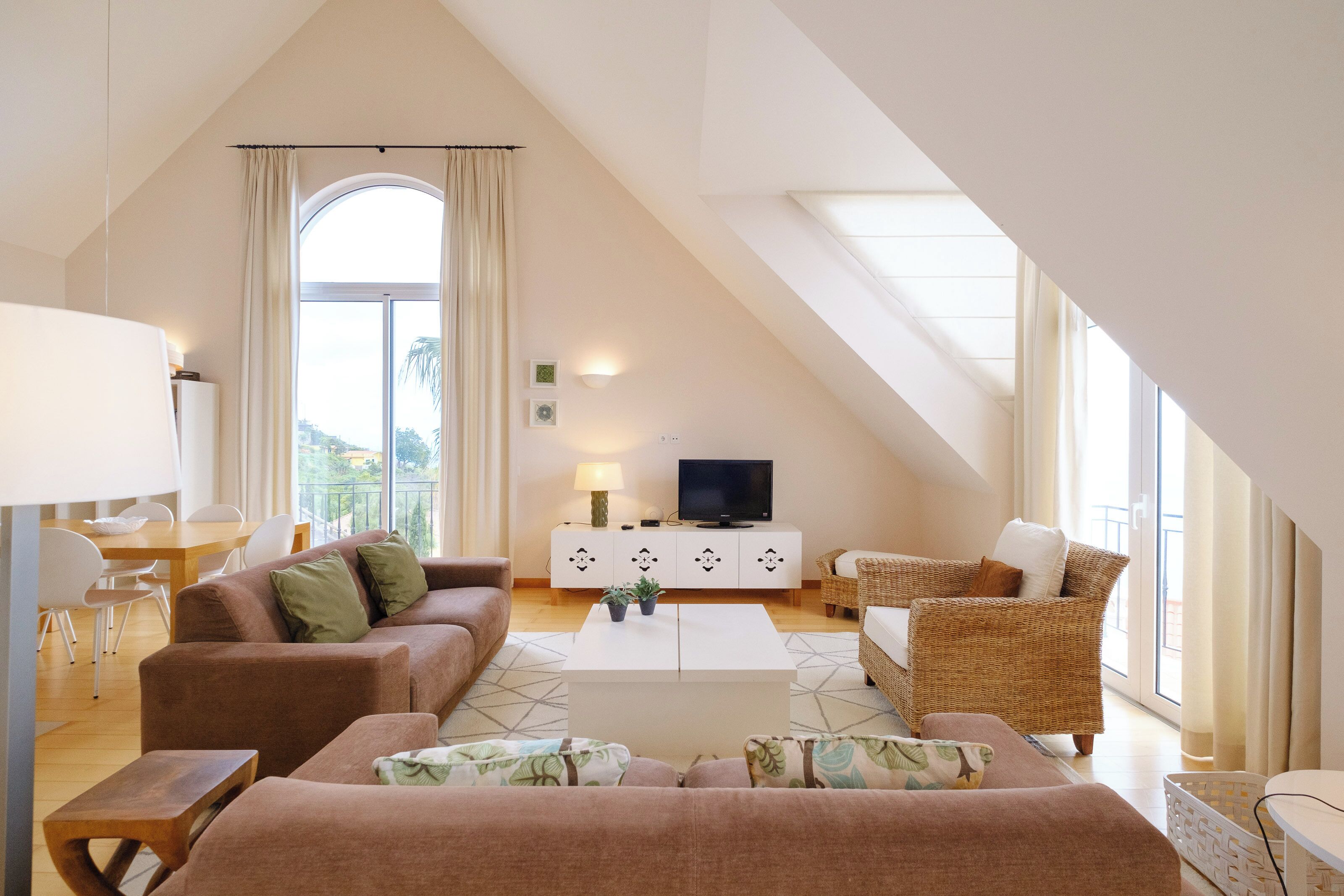 A spacious living room with a high ceiling, a large window, a white coffee table, and a television on a white cabinet.