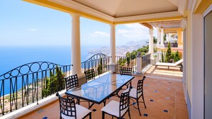 A balcony with a glass table and chairs, overlooking a scenic view of the sea and mountains.